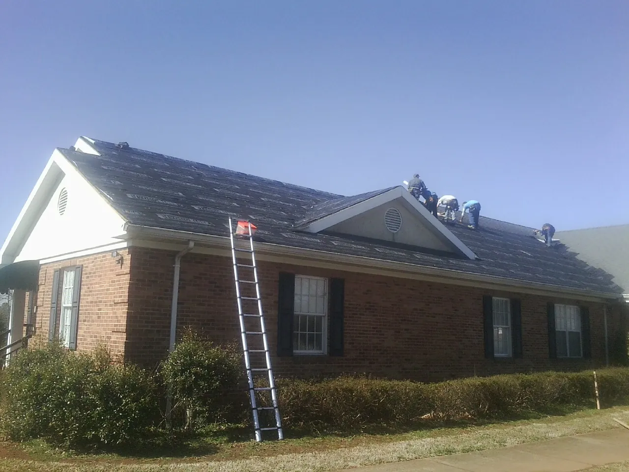 Crew tearing shingles off a brick ranch roof with a ladder set — Emergency Roof Repair work by our crew in Hewitt